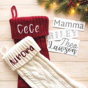 May include: A deep red knitted Christmas stocking with the white embroidered name CeCe and a cream-colored knitted stocking with the dark red embroidered name Aurora. Three white rectangular labels display the names Mamma, RILEY, and Theo Lawson.