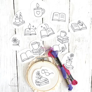 May include: A collection of black and white line art stickers featuring books, candles, mugs, and flowers. Embroidery floss in purple and pink hues sits near an embroidery hoop with a book and glasses design. The items are arranged on a white wooden surface.