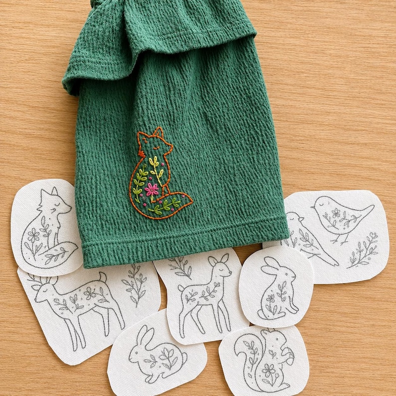 May include: A dark green textured fabric piece featuring an orange embroidered fox with floral details, surrounded by several white fabric patches with black line art illustrations of a fox, deer, rabbit, squirrel, and bird with floral accents.