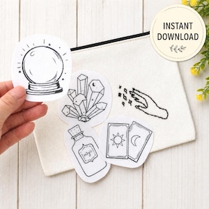 May include: A white canvas pouch with a black zipper, featuring several black and white embroidered patches. Patches include a crystal ball, crystals, a hand with stars, tarot cards, and a potion bottle with the word "magic".