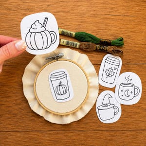 May include: A wooden embroidery hoop containing off-white fabric with a paper cutout of a pumpkin-shaped mug. Additional paper cutouts feature a coffee cup with a maple leaf, a mug with a moon and stars, and a mug with a witch hat. Two skeins of green and brown embroidery floss are included.