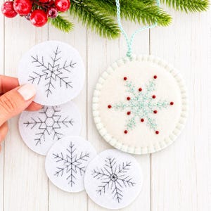 May include: A set of circular white felt ornaments featuring black embroidered snowflake patterns. One larger ornament is finished with a light blue braided hanging cord, light blue stitching, and small red beads accenting the snowflake.