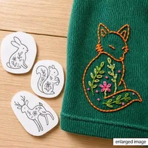 May include: A dark green fabric featuring an orange embroidered fox outline filled with green leaves and pink flowers, alongside three white paper patches displaying black line drawings of a rabbit, a squirrel, and a deer with floral details.