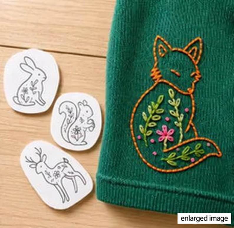 May include: A dark green fabric featuring an orange embroidered fox outline filled with green leaves and pink flowers, alongside three white paper patches displaying black line drawings of a rabbit, a squirrel, and a deer with floral details.