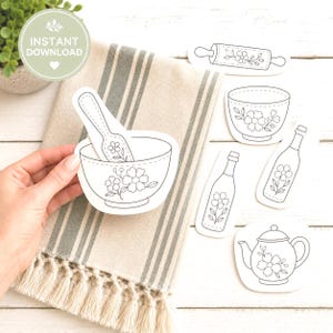 May include: A set of black and white line art stickers featuring kitchenware including a bowl with a spoon, a rolling pin, a tea cup, two bottles, and a teapot. Each item is decorated with a floral pattern. The stickers are displayed on a beige towel.