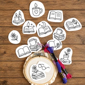 May include: A collection of white paper cutouts featuring black line art illustrations of books, mugs, candles, flowers, and glasses, arranged around a wooden embroidery hoop containing fabric and two skeins of purple and pink embroidery floss.