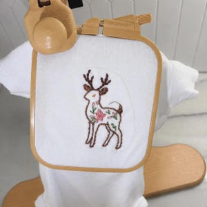 May include: A white fabric square held in a light brown plastic Nurge embroidery hoop. The fabric features a hand-stitched embroidery design of a deer with brown outlines and pink and green floral details on its body, centered on the white cloth.
