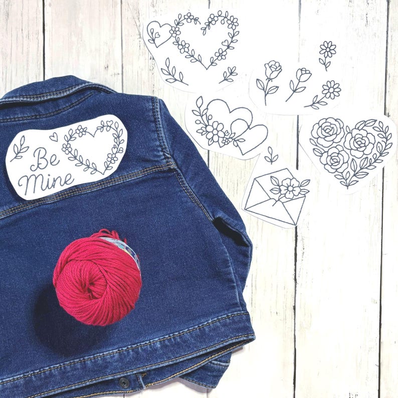 May include: A dark blue denim jacket with a red ball of yarn and several white paper cutouts featuring black line drawings of hearts, flowers, and an envelope. One cutout says "Be Mine".