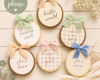 May include: Six circular wooden embroidery hoops containing fabric with embroidered phrases including hello world, heaven sent, it's a boy, she's here, it's a girl, and he's here. Each hoop is decorated with a frayed fabric bow in various colors.