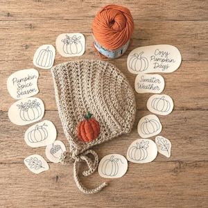 May include: A beige crocheted bonnet featuring a small orange pumpkin appliqué with a green stem. The bonnet has matching ties. Surrounding the bonnet are paper cutouts with black ink drawings of pumpkins, leaves, an acorn, and text.