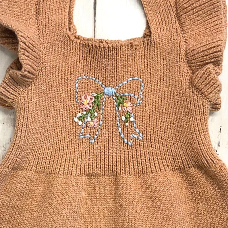 May include: A tan ribbed knit garment featuring ruffled sleeves and a central embroidered design of a light blue bow accented with small pink and yellow flowers and green leaves. The fabric has a textured, vertical ribbed pattern throughout.
