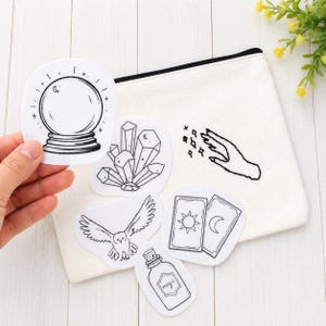 May include: A collection of black and white embroidered patches with mystical and magical themes. Patches include a crystal ball, crystals, an owl, tarot cards, a potion bottle with the word "magic", and a hand with stars. The patches are on a white canvas bag.