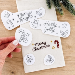 May include: A white fabric towel featuring embroidered Christmas designs including the text Merry Christmas, Merry and Bright, and Holly Jolly, alongside various patches depicting candy canes, bells, holly, stars, and a snowflake.