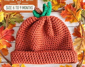 Pode incluir: Um gorro de crochê em tom laranja queimado com um design texturizado de abóbora e um caule verde no topo. O gorro possui uma aba dobrada e contém o texto READY TO SHIP e SIZE 6 TO 9 MONTHS dentro de uma caixa retangular marrom.
