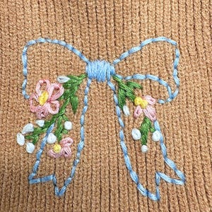 May include: A tan ribbed knit fabric featuring an embroidered light blue bow outline. The bow is decorated with green stems, small white buds, and pink flowers with yellow centers, creating a detailed floral pattern on the textured material.