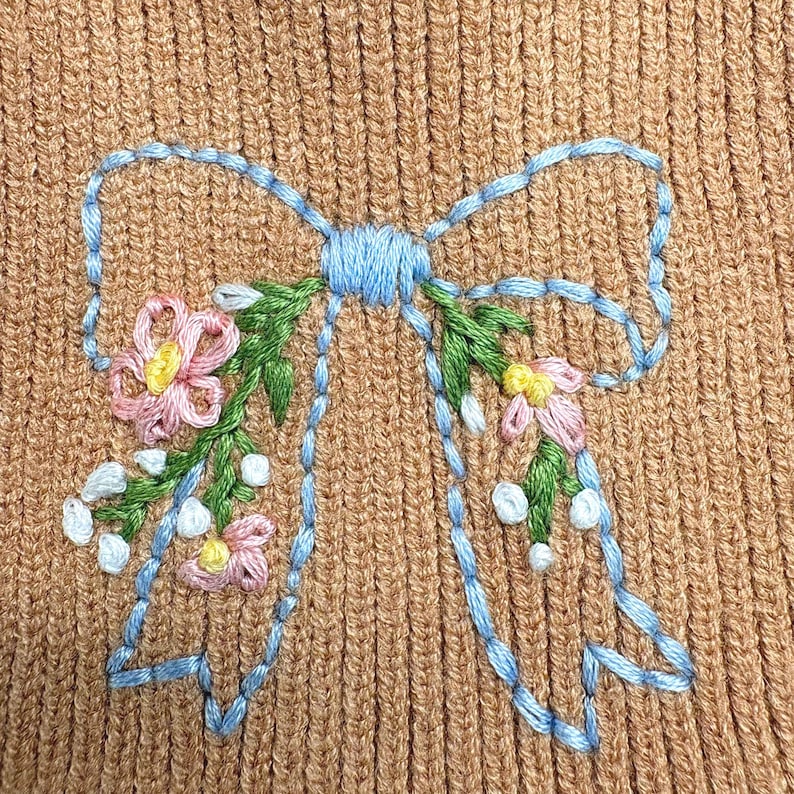 May include: A tan ribbed knit fabric featuring an embroidered light blue bow outline. The bow is decorated with green stems, small white buds, and pink flowers with yellow centers, creating a detailed floral pattern on the textured material.