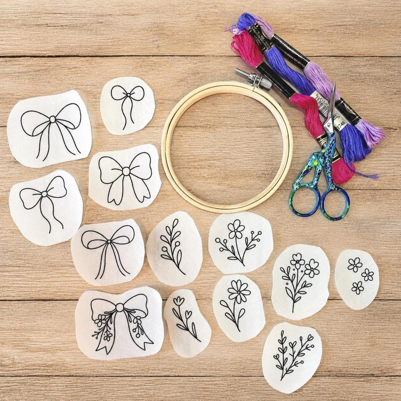 May include: An embroidery kit featuring a wooden hoop, three skeins of embroidery floss in pink, purple, and light purple, a pair of patterned scissors, and several white fabric patches with black line art designs of bows and floral sprigs.