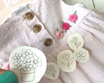 May include: A light pink baby dress with embroidered pink roses and green leaves near the collar. The dress has three brown buttons and a white tag. Several small, white, oval-shaped patches with flower designs are also present.