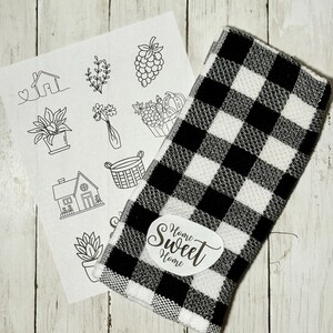 May include: A black and white checkered kitchen towel with a white oval label that reads "Home Sweet Home" in script. A sheet of paper with line drawings of houses, plants, and other home-themed images is next to the towel.