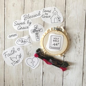 May include: An assortment of embroidery supplies, including a hoop with a "Holy Bible" design, several paper cutouts with religious phrases and symbols, and a collection of colorful embroidery floss. The items are arranged on a light wood-grain surface.