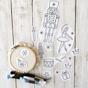 May include: A collection of Christmas-themed embroidery patterns and supplies. Includes a wooden embroidery hoop with a snowflake and ornament design, along with various cut-out designs of a nutcracker, ballerina, and other festive elements. Embroidery floss in shades of blue and white.