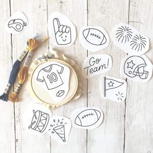 May include: An embroidery hoop with a jersey design, football-themed cutouts, and spools of thread. The cutouts include a football, a helmet, a flag, and the words "Go Team!" on a light wood grain surface.
