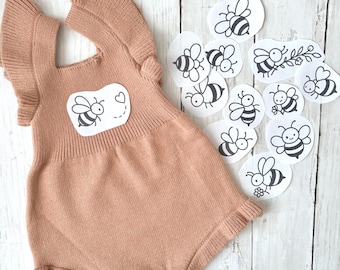 May include: A light brown knitted romper with frilled sleeves and a bee graphic on the front. Several bee stickers are scattered around the romper on a white wooden surface. The romper has a heart-shaped detail.