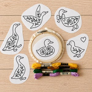 May include: A set of six white fabric patches featuring black line art of ducks with floral patterns, one duck inside a wooden embroidery hoop, and four skeins of embroidery floss in yellow, brown, green, and purple colors for needlework.