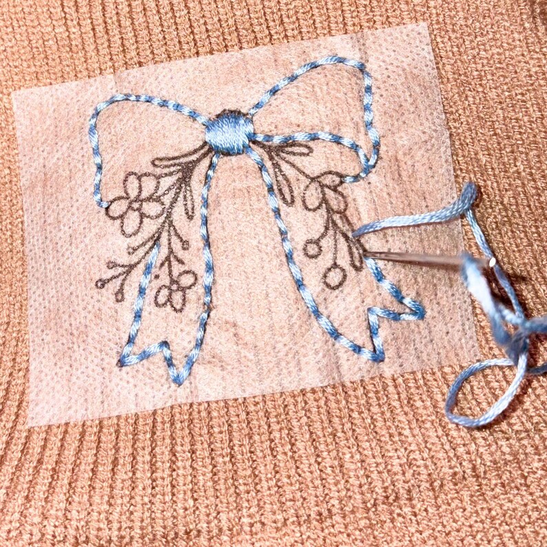 May include: A tan knit fabric featuring a translucent stabilizer sheet with a printed black line drawing of a bow and floral sprigs. A needle with light blue embroidery thread is partially stitched along the outline of the bow shape.