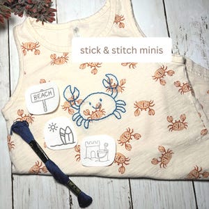 May include: A cream-colored sleeveless tank top featuring a repeating pattern of small orange crabs. A blue embroidered crab sits in the center. Three white stick and stitch mini patches show a beach sign, surfboards, and a sandcastle. A skein of dark blue embroidery floss is included.