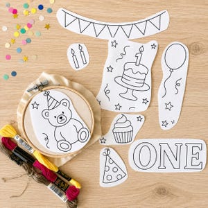 May include: A set of black and white line art paper cutouts for embroidery featuring a teddy bear in an embroidery hoop, a birthday cake with a candle, a balloon, a cupcake, a party hat, a banner, and the word ONE, with colorful embroidery floss.