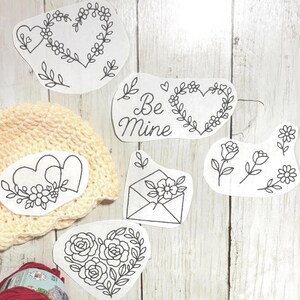 May include: Various black line art designs on white paper, featuring floral hearts, roses, and the text "Be Mine." A cream-coloured crocheted hat and a ball of red yarn are also present.