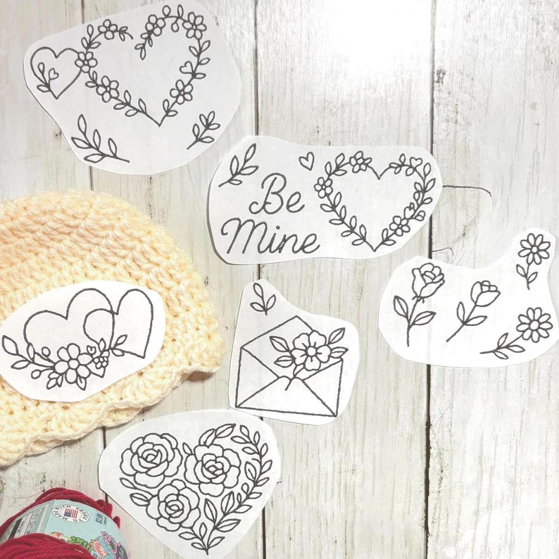 May include: Various black line art designs on white paper, featuring floral hearts, roses, and the text "Be Mine." A cream-coloured crocheted hat and a ball of red yarn are also present.