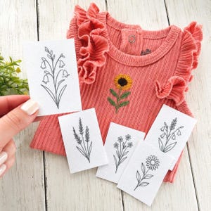 May include: A coral-colored ribbed sleeveless top with ruffled shoulders and a small embroidered sunflower on the chest. The top is displayed with five white cards, each featuring a distinct black line drawing of a different wildflower.