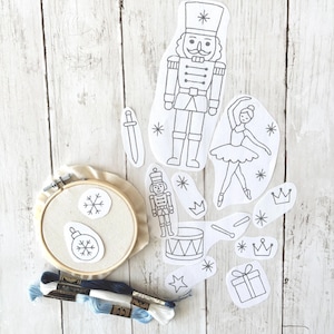 Könnte beinhalten: Stick-Set mit einem Holzreifen, Stoff und verschiedenen ausgeschnittenen Formen, darunter ein Nussknacker, eine Ballerina und Schneeflocken. Blau-weißes Stickgarn ist ebenfalls enthalten, ideal für ein weihnachtliches Bastelprojekt.