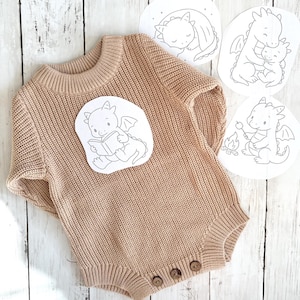 May include: A beige knitted baby romper with short sleeves and wooden buttons. A white sticker featuring a cartoon dragon reading a book is on the front. Three other dragon illustrations are also visible.