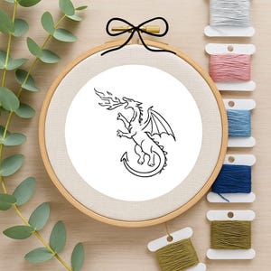 May include: A round wooden embroidery hoop containing white fabric with a black line art illustration of a fire-breathing dragon. The hoop features a black bow at the top, and a collection of six colorful embroidery thread spools is arranged nearby.