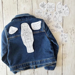 Könnte beinhalten: Dunkelblaue Jeansjacke mit mehreren weißen, umrissartigen Weihnachtspatches. Zu den Patches gehören ein Nussknacker, eine Trommel, Stechpalmen, Ornamente und Schleifen. Die Jacke liegt auf einer hellen Holzmaserung.