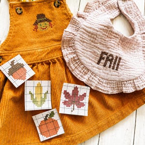 May include: A mustard yellow corduroy overall dress featuring a cross-stitched scarecrow, a ruffled light pink muslin bib with the embroidered text "FALL", and four square paper patterns depicting an acorn, corn, a maple leaf, and a pumpkin.