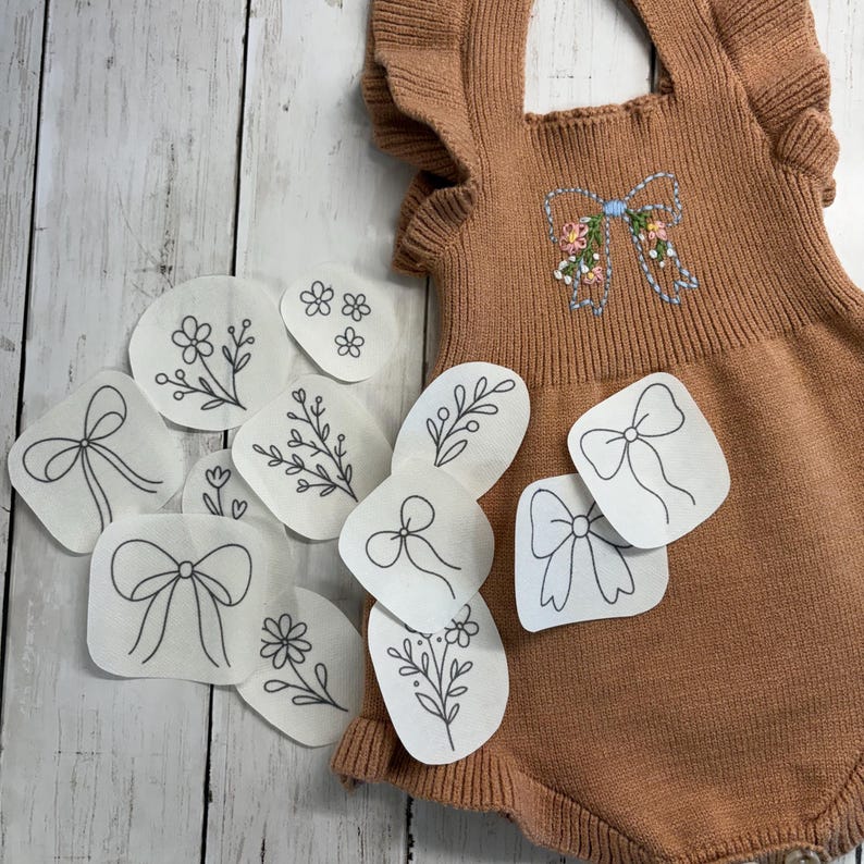 May include: A tan knit romper with ruffled sleeves and a central embroidered bow featuring small floral accents, accompanied by several white adhesive patches displaying black line art illustrations of bows and various flowers.