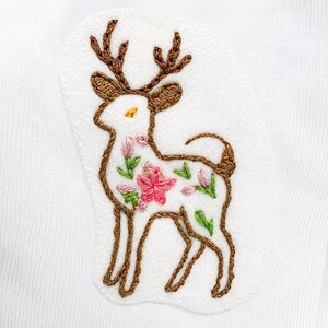 May include: An embroidered patch on a white fabric background featuring a deer outline in brown thread. The deer has brown antlers and is filled with small, colorful floral embroidery in shades of pink, green, and yellow, creating a detailed design.