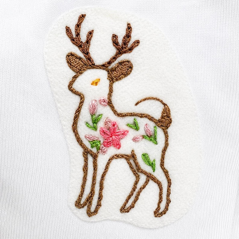 May include: An embroidered patch on a white fabric background featuring a deer outline in brown thread. The deer has brown antlers and is filled with small, colorful floral embroidery in shades of pink, green, and yellow, creating a detailed design.