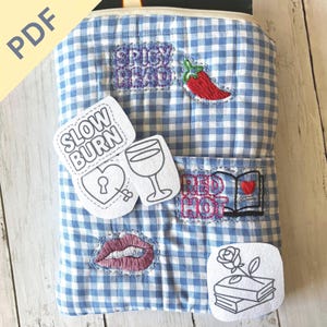 Peut inclure: Une housse de livre en tissu vichy bleu et blanc avec des patchs brodés. Les patchs incluent un piment rouge, une paire de lèvres, un verre à vin et les phrases "Spicy Head", "Slow Burn" et "Red Hot".