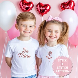 May include: Two children wearing white t-shirts with Valentine's Day designs. One shirt says "Be Mine" in red script, the other has a red rose. Pink balloons and heart-shaped balloons are in the background.