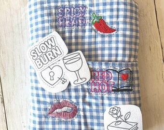 May include: A light blue and white gingham quilted book sleeve featuring embroidered patches of a red chili pepper, a heart with a keyhole, a wine glass, a pair of lips, and an open book with a red heart. Text includes SPICY READ, SLOW BURN, and RED HOT.