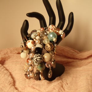 May include: A charm bracelet featuring a variety of beads in different shapes, sizes, and textures, including white, black, brown, and blue colours with patterns like zebra stripes, leopard spots, and polka dots, displayed on a black hand stand.