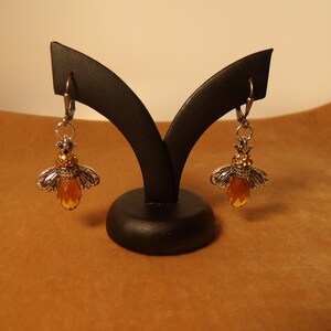 May include: A pair of bee-shaped earrings. The earrings have silver-coloured wings, a golden-coloured body, and a faceted, amber-coloured crystal. The earrings are displayed on a black jewellery stand against a brown background.