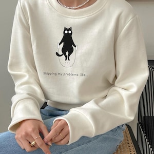 Könnte beinhalten: Cremefarbenes Sweatshirt mit einer Illustration einer schwarzen Katze, die Seil springt. Der Text "Skipping my problems like..." ist unter der Katze aufgedruckt. Das Sweatshirt hat lange Ärmel und einen Rundhalsausschnitt.
