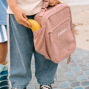 May include: A pink corduroy lunch bag with the name "EMMA" printed on the front. A banana is being placed in the side pocket. The bag has a top handle and a zipper closure. The person is wearing blue jeans and black and white sneakers.