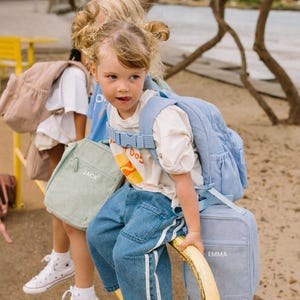 May include: Two young children with backpacks and lunch bags. One child wears a light blue backpack and matching lunch bag with the name "Emma". The other child has a light green lunch bag with the name "Jack". Both children are wearing sneakers.